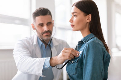 Lady coming to clinic for heart and lungs checkup, male doctor using stethoscope, listening to female patient's breath or heartbeat, sitting in clinic office
